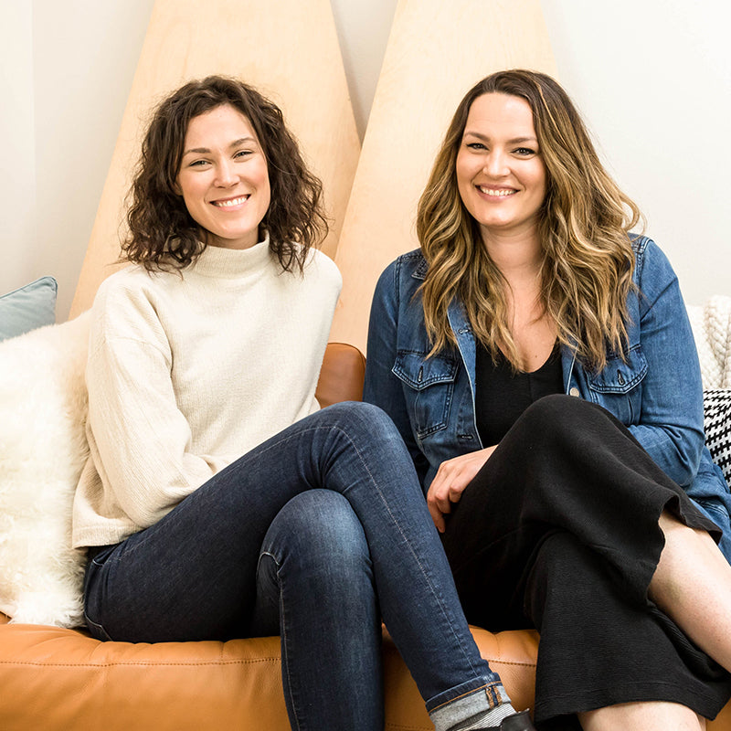 Two women sitting on a leather couch smiling in a bright, cozy room