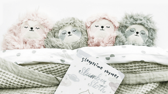 Four plush sloth toys tucked into a polka-dot blanket beside a children’s book titled Sleepytime Rhymes, cozy bedtime scene