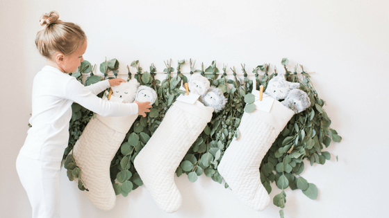 Girl placing plush Slumberkins toys into cream quilted stockings hung on eucalyptus garland against a white wall