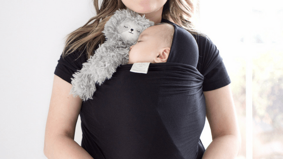 Parent wearing a black Solly baby wrap holding a sleeping infant with a gray Slumberkins plush tucked by the baby’s head