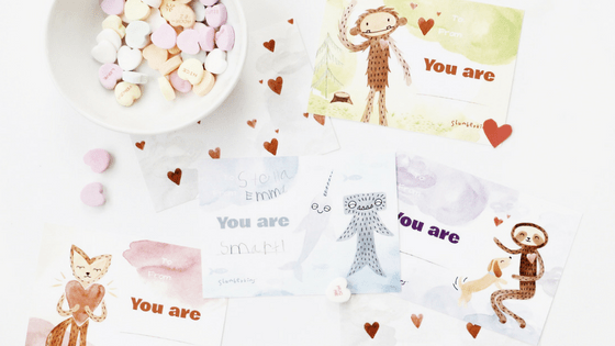Flat lay of printable “You are” Valentine’s cards with heart stickers and a bowl of conversation-heart candies on a white background