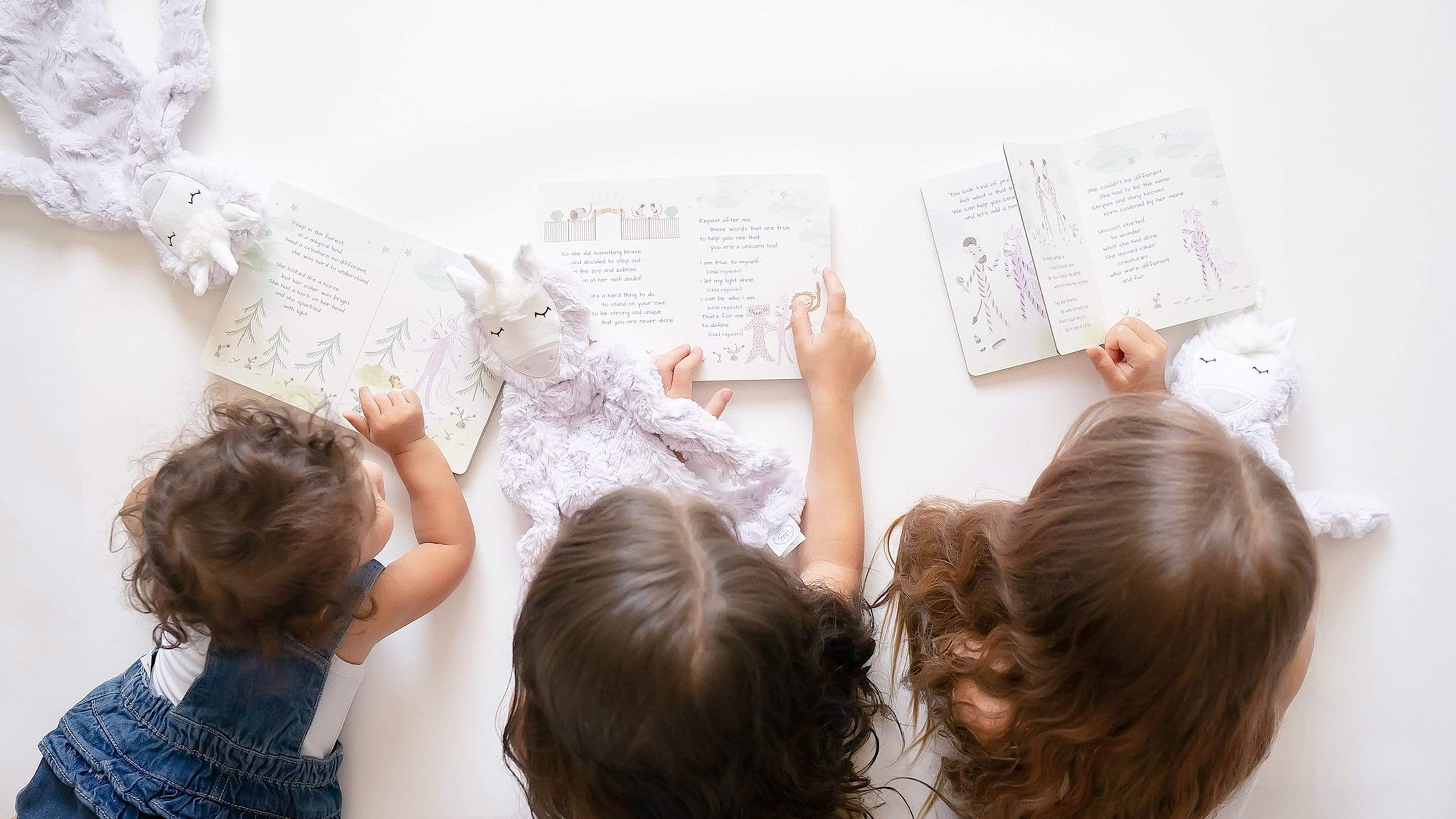 Three children lying on a white surface reading board books with lavender unicorn lovey plushes