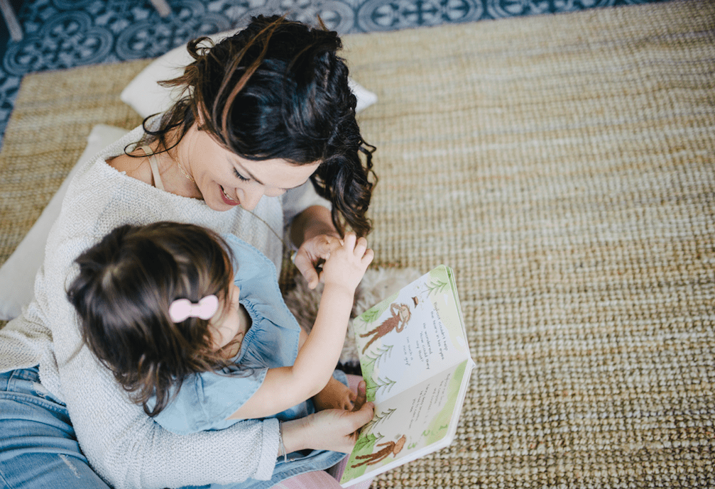 Woman reading a picture book with a toddler on her lap, both smiling on a woven rug