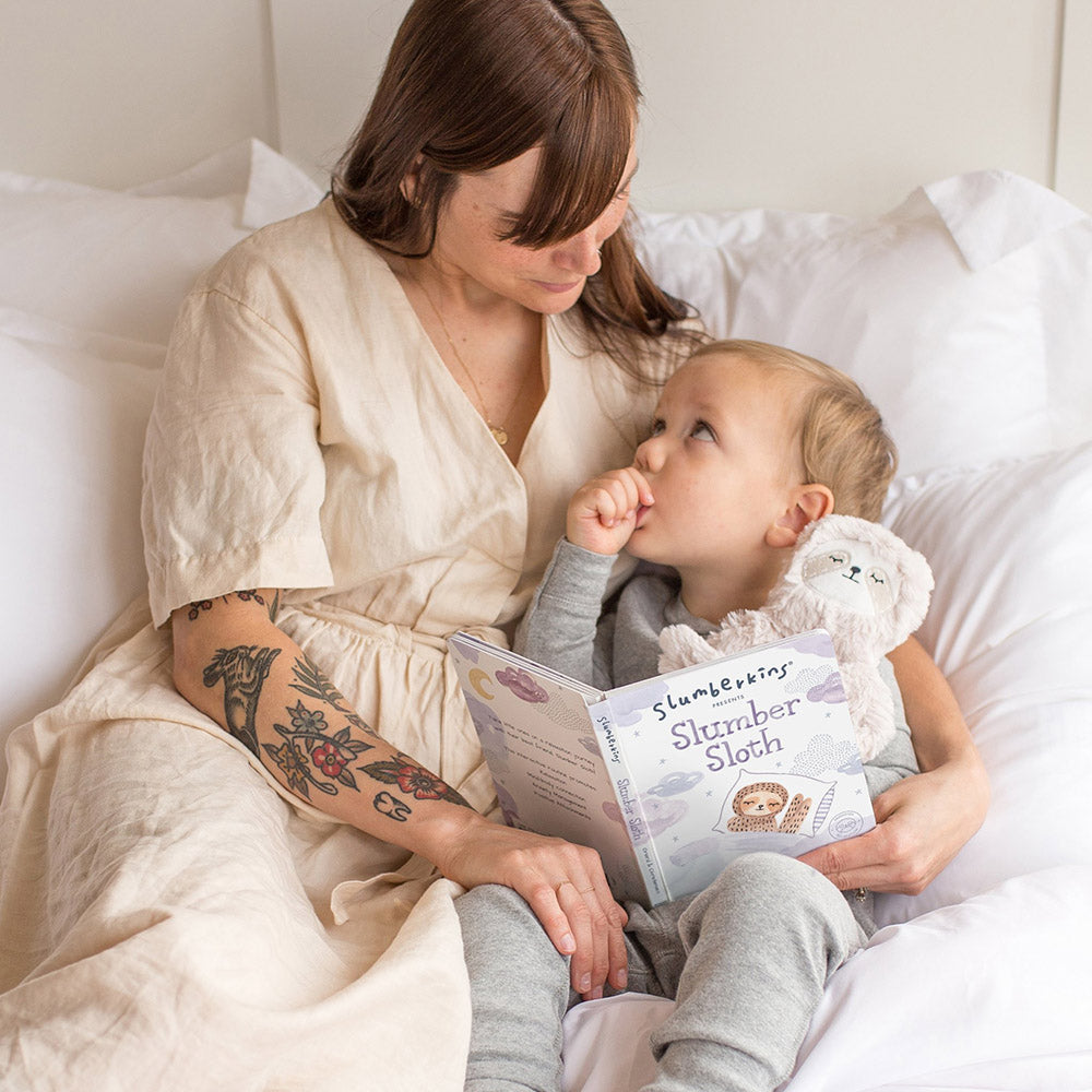 Woman reading the book Slumber Sloth to a toddler holding a sloth plush on a white bed