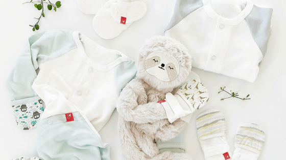 Sloth plush hugging baby mittens arranged with pastel onesies and socks on a white background