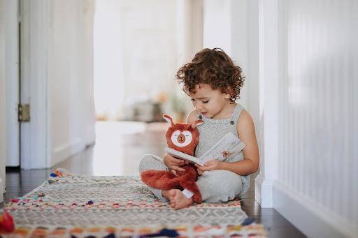 Toddler sitting on a patterned rug reading a book while holding a rust-colored plush alpaca in a bright hallway