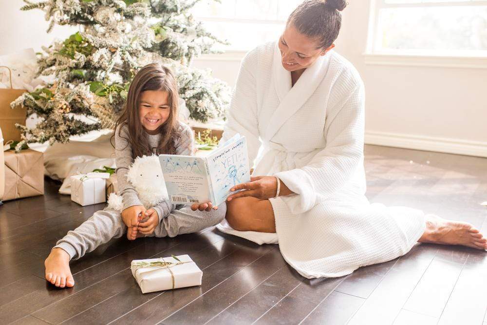 Mother reading a Slumberkins book to a smiling child by a decorated Christmas tree, child holding a white plush and wrapped gift
