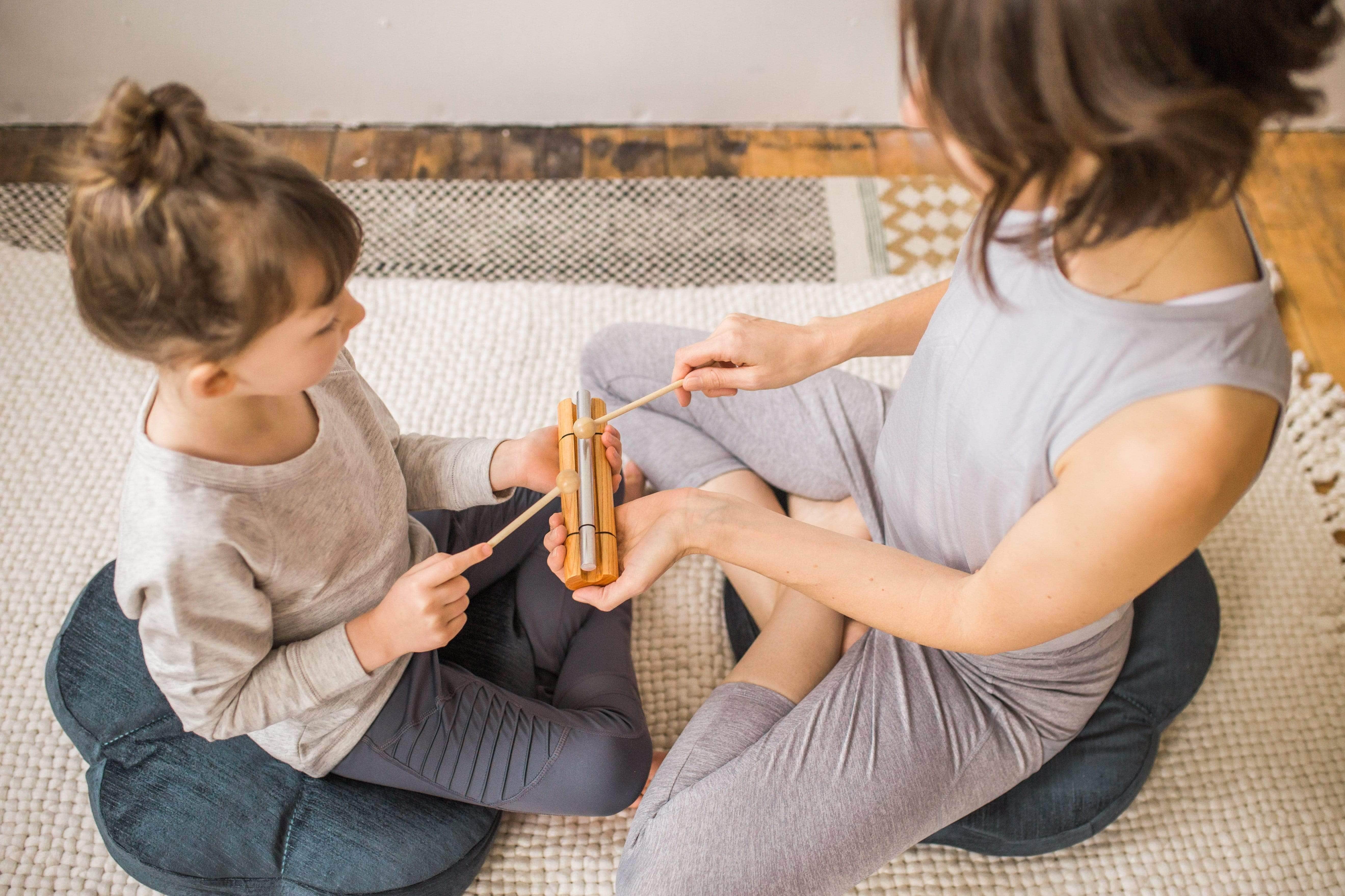 Adult and child gently tapping a wooden chime together while seated on floor cushions for a mindful moment