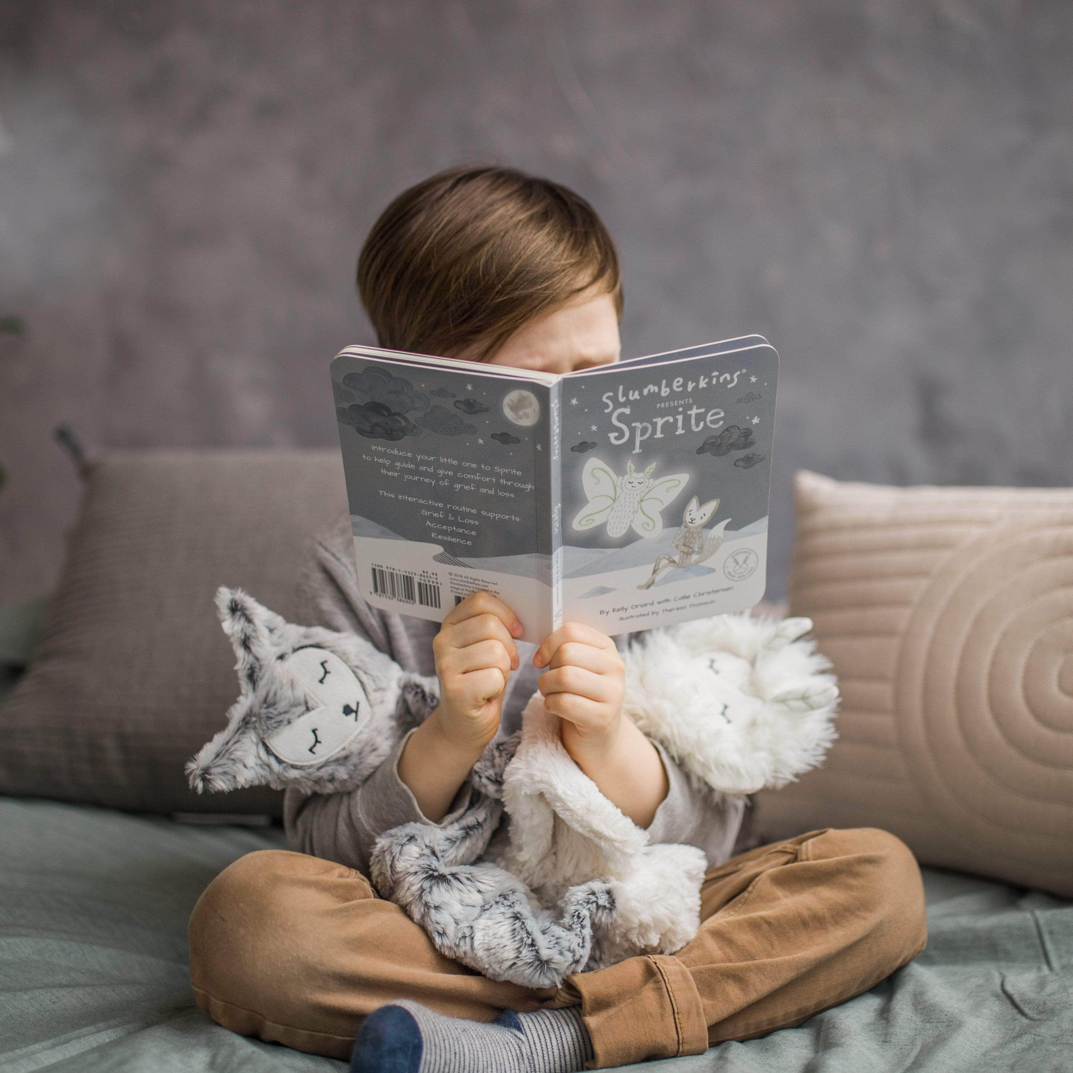 Child reading Slumberkins Sprite board book while hugging two plush animals on a couch