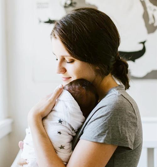 Mother holding swaddled newborn against her chest in a softly lit nursery, eyes closed in a peaceful embrace