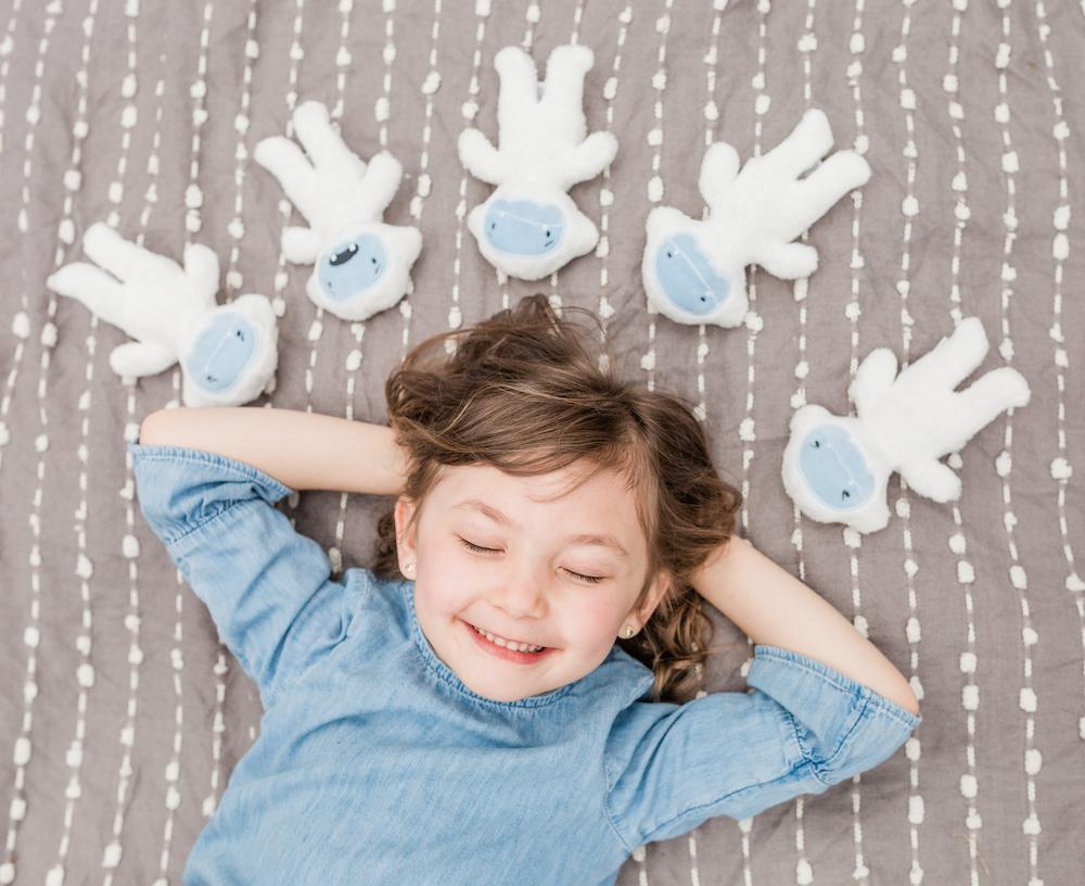 Girl lying on gray blanket, smiling, eyes closed, arms behind her head, surrounded by five white plush toys with blue faces