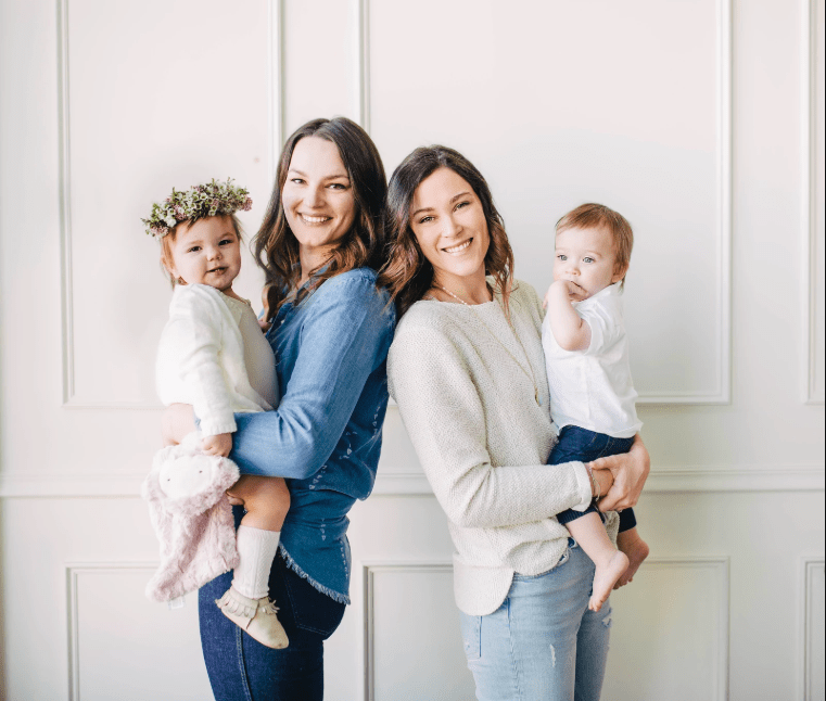 Two smiling mothers holding babies in a bright studio against a white paneled wall