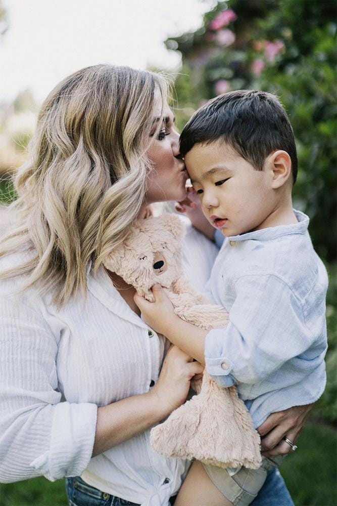 Mother kissing young boy on forehead while holding a plush honey bear in a sunlit garden