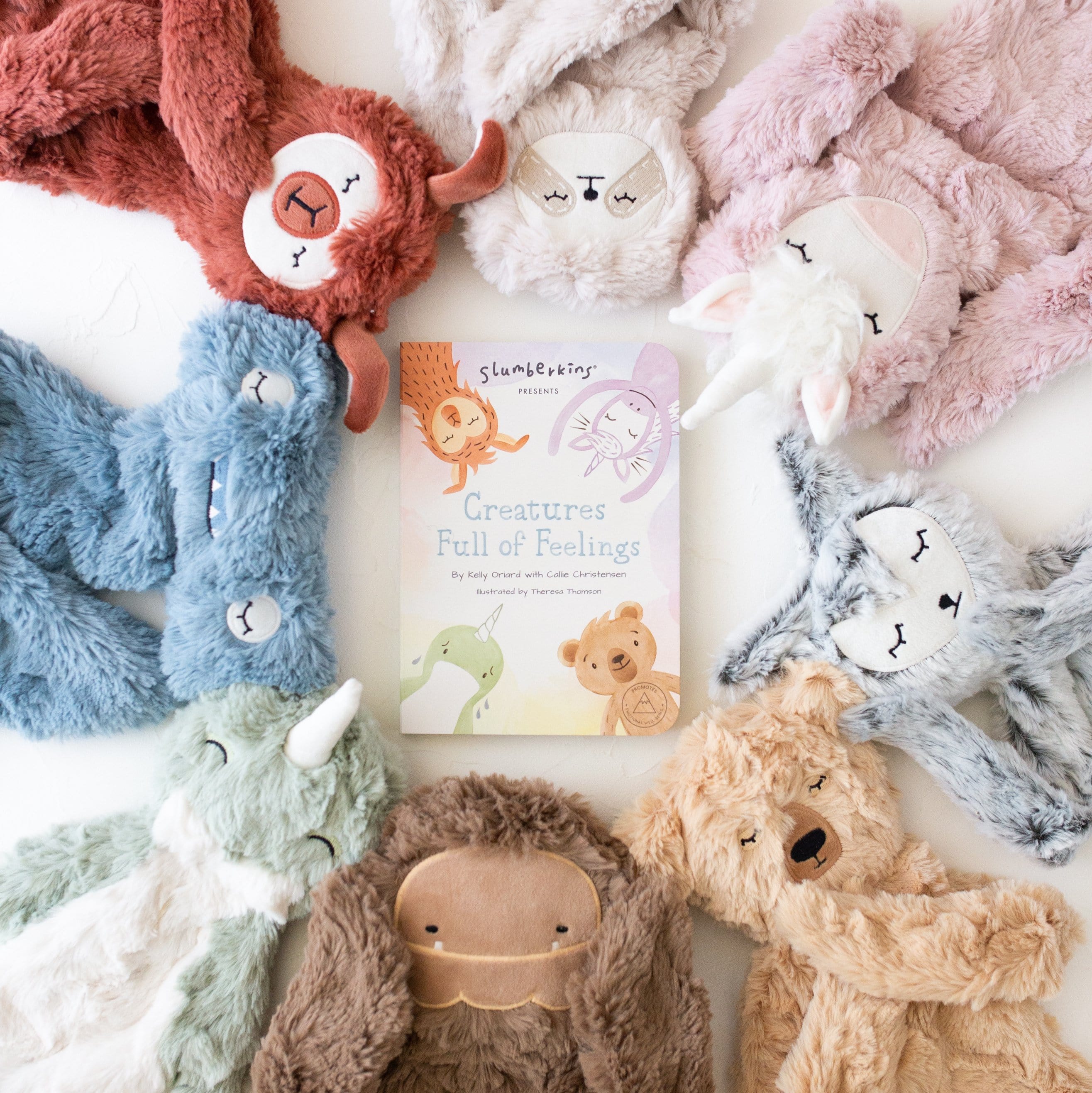 Flat lay of Creatures Full of Feelings book with pastel plush loveys showing soft fur and sleepy faces on white background