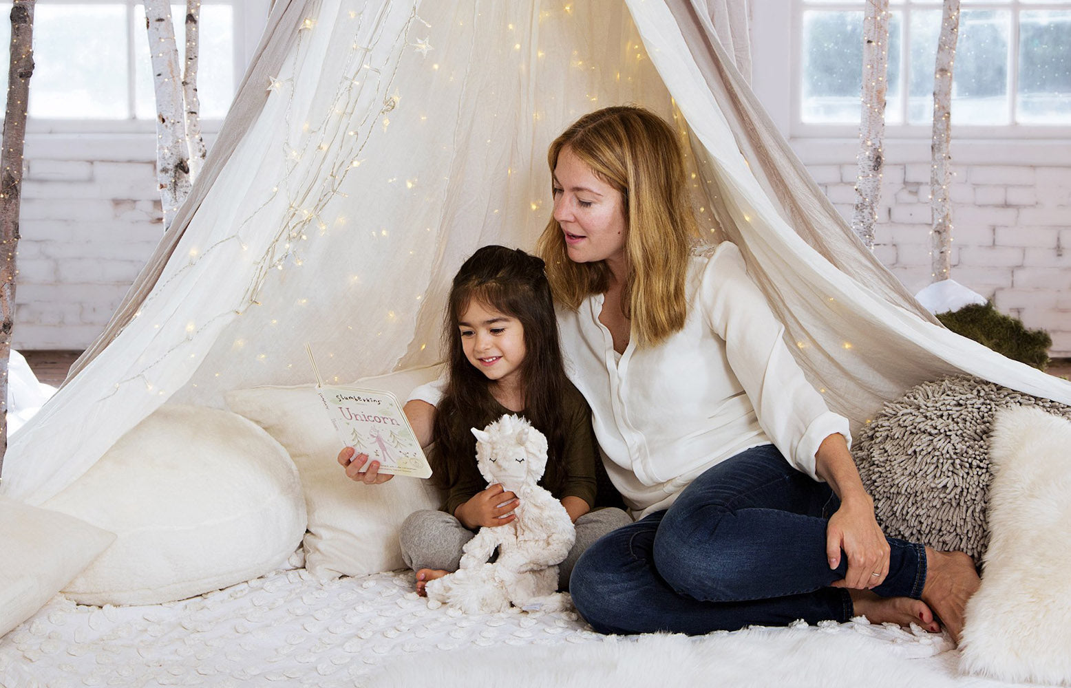 Mother reading a unicorn book to her daughter inside a cozy canopy tent with fairy lights, child cuddling a plush unicorn