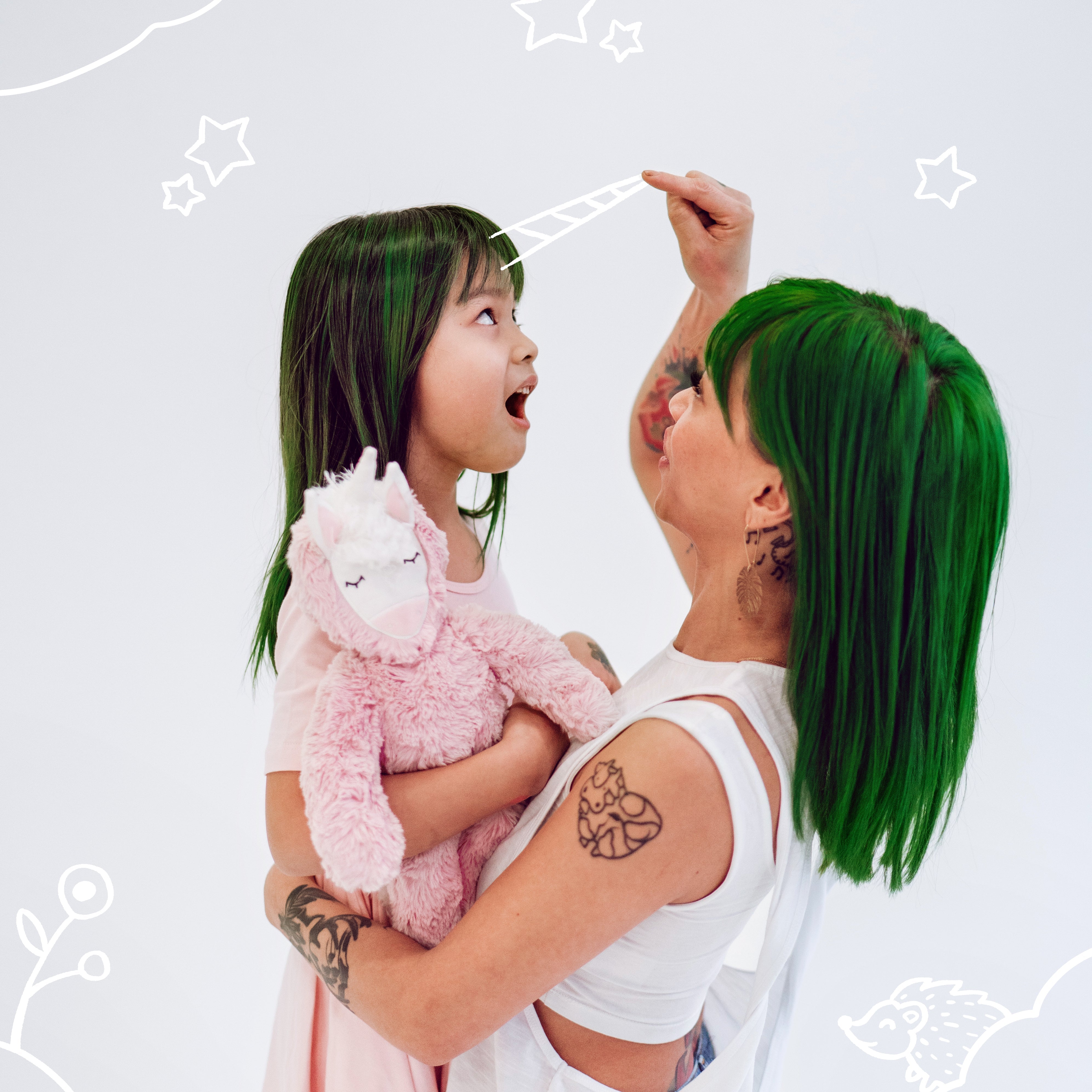 Adult holding a child with a pink plush unicorn as the adult draws a doodle unicorn horn on her forehead, white background