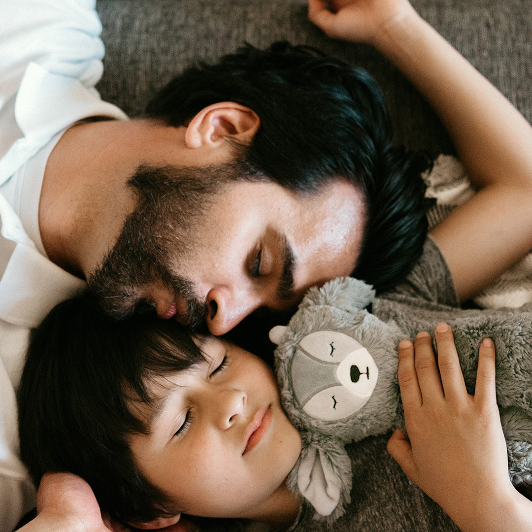 Man and child lying together, peacefully cuddling a gray plush sloth while resting close