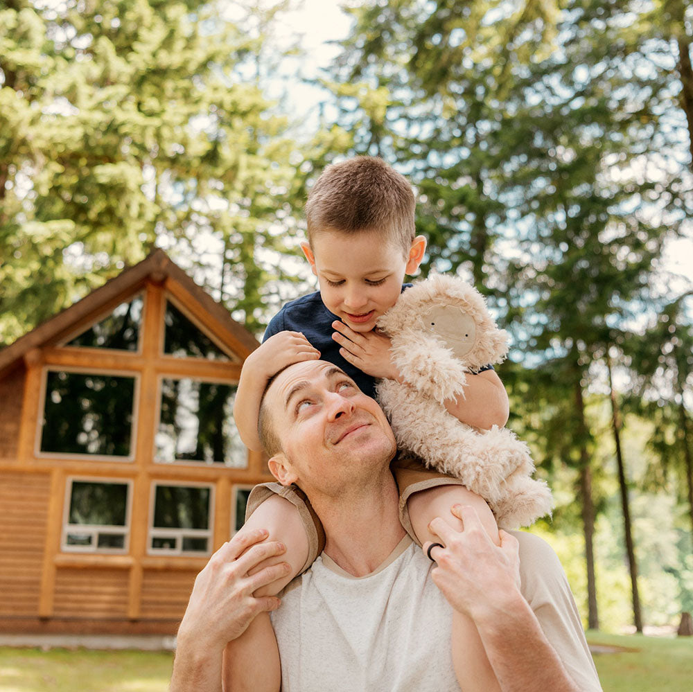 Father carrying young son on his shoulders while the boy hugs a fluffy plush toy in front of a wooden cabin in the woods