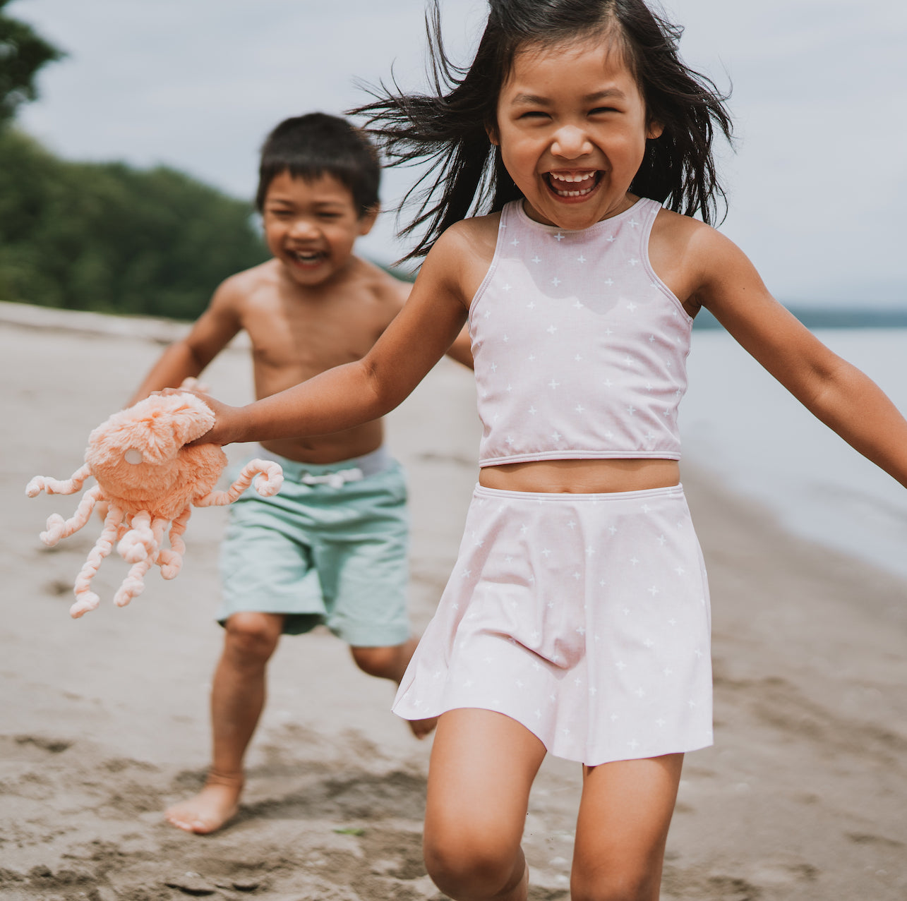 Young girl laughing and running on a beach while a younger boy chases her, girl holding a plush pink octopus