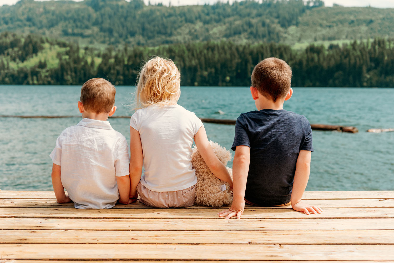 Three children sitting on a wooden dock by a tree-lined lake, facing the water while the middle child holds a teddy bear