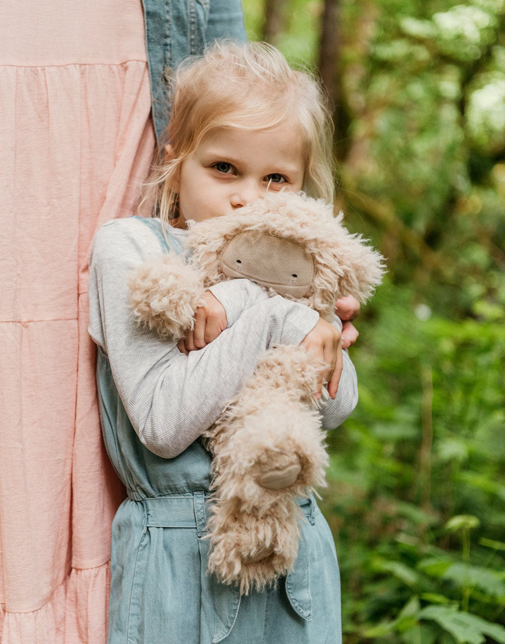 Little girl hugging a fluffy beige plush Slumberkins toy while standing outdoors next to an adult in a pink dress