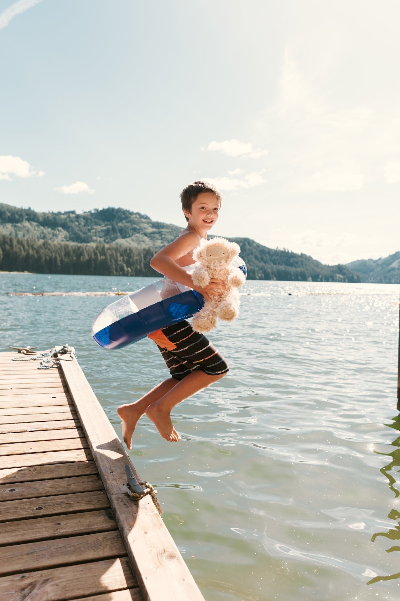 Boy jumping from a wooden dock into a lake holding a plush toy and wearing a blue inflatable swim ring with forested hills behind