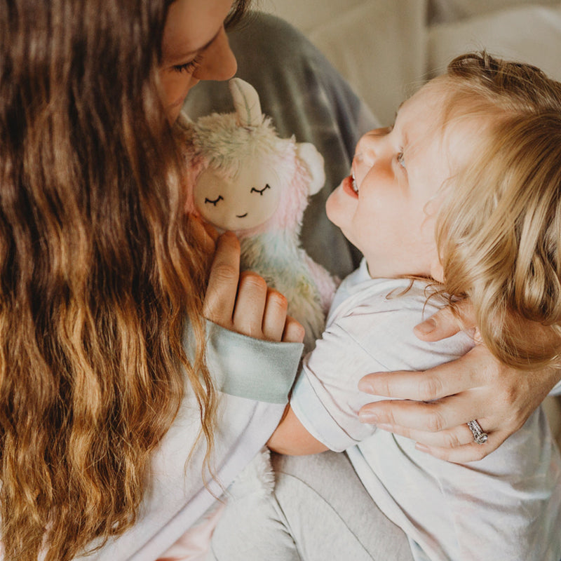 Adult cuddling a smiling toddler while holding a pastel rainbow unicorn plush toy