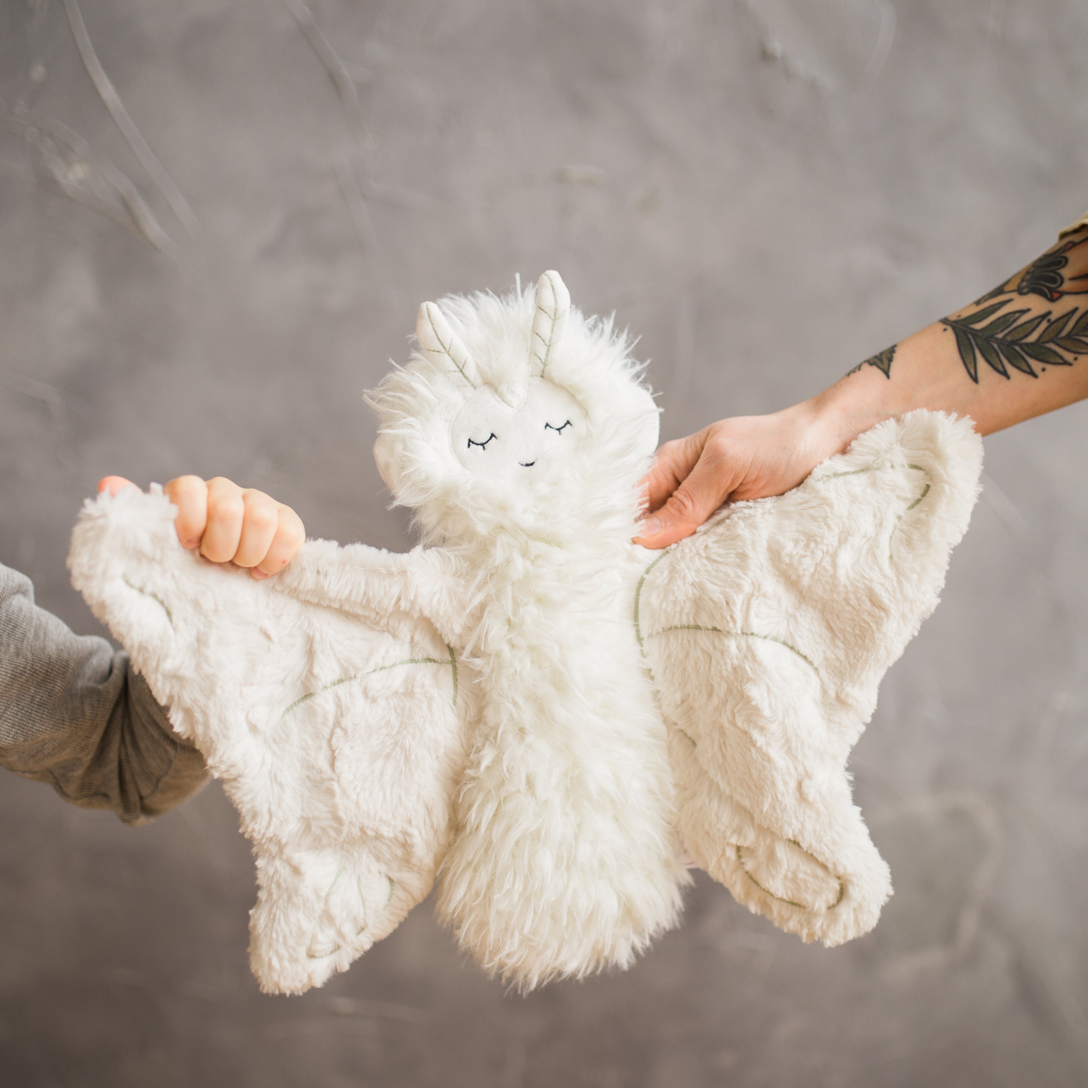 White plush unicorn lovey with fluffy fur and embroidered closed eyes, held by adult and child against gray background