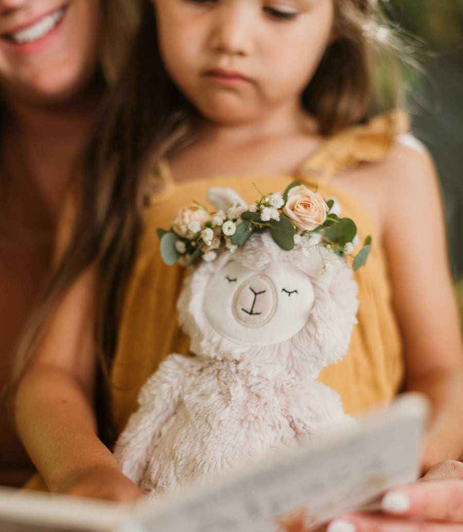 Young girl and caregiver reading a book together while the child holds a plush llama wearing a flower crown
