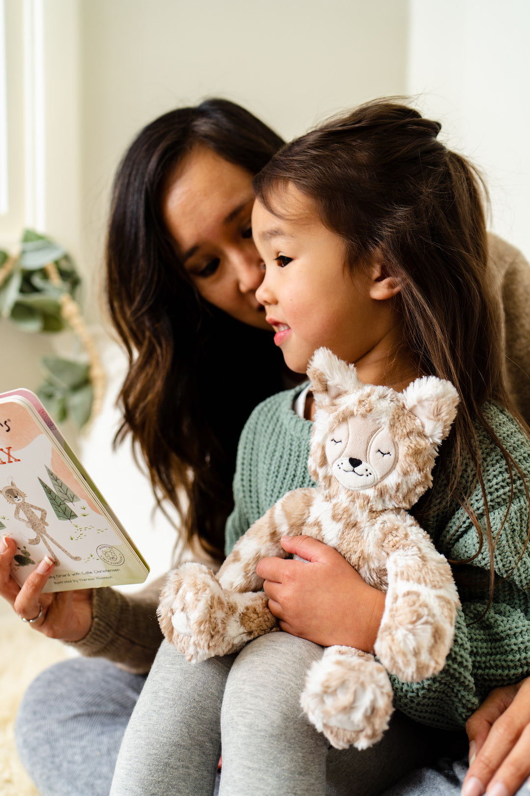 Mother reading a board book to a young girl who holds a beige spotted plush fox on her lap in a bright cozy room