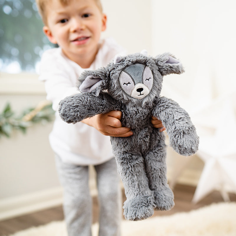 Child offering a gray plush animal with embroidered closed eyes toward the camera in a bright bedroom setting