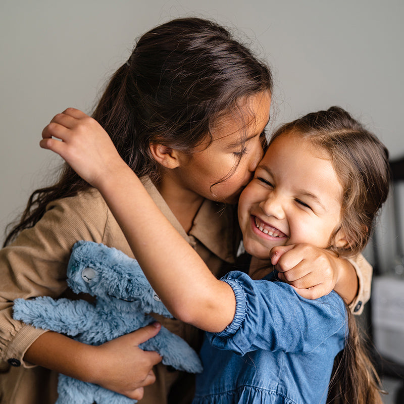 Two young girls hugging as one kisses the other’s cheek while the other laughs, older child holding a blue plush rabbit in a warm indoor setting