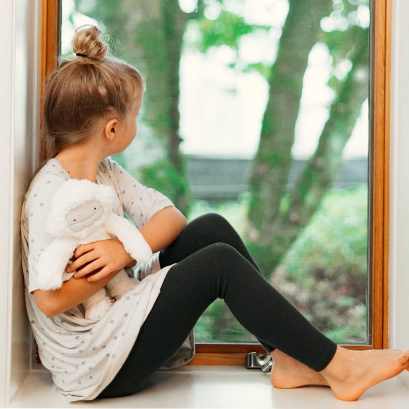 Young girl sitting on a window seat hugging a white plush toy and looking out at the trees