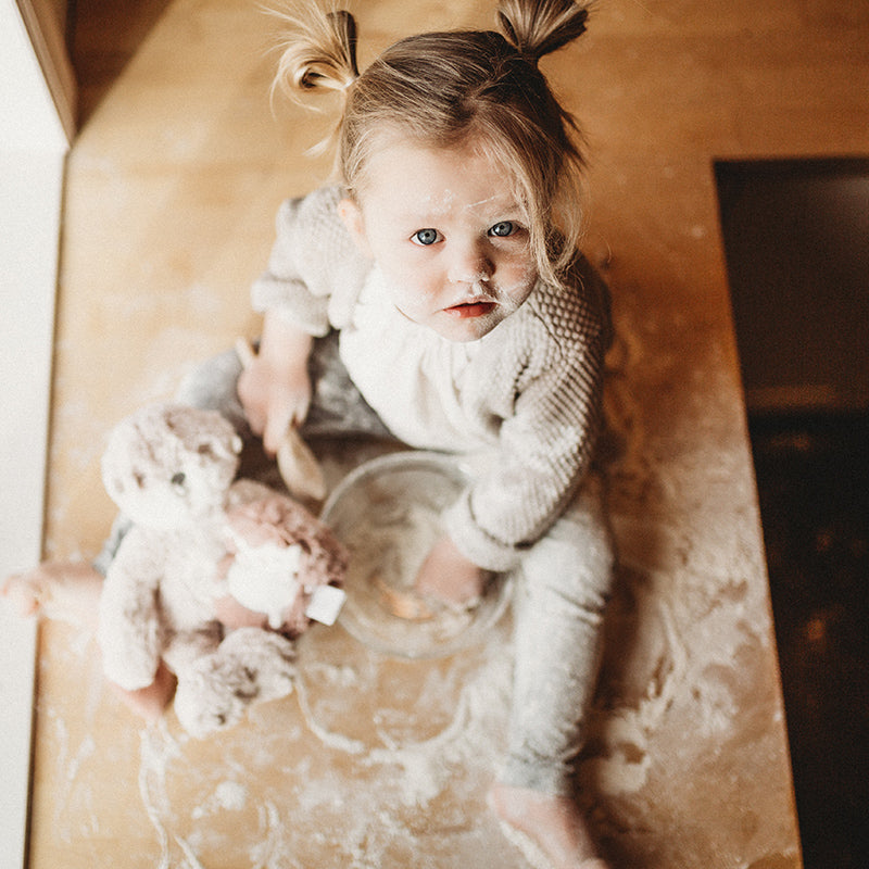 Toddler sitting on a flour-covered kitchen counter mixing batter with a wooden spoon while a plush stuffed animal sits beside them