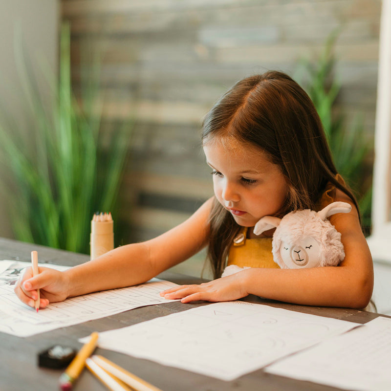 Young girl writing at a table holding a plush lamb, concentrating in a cozy classroom-like setting
