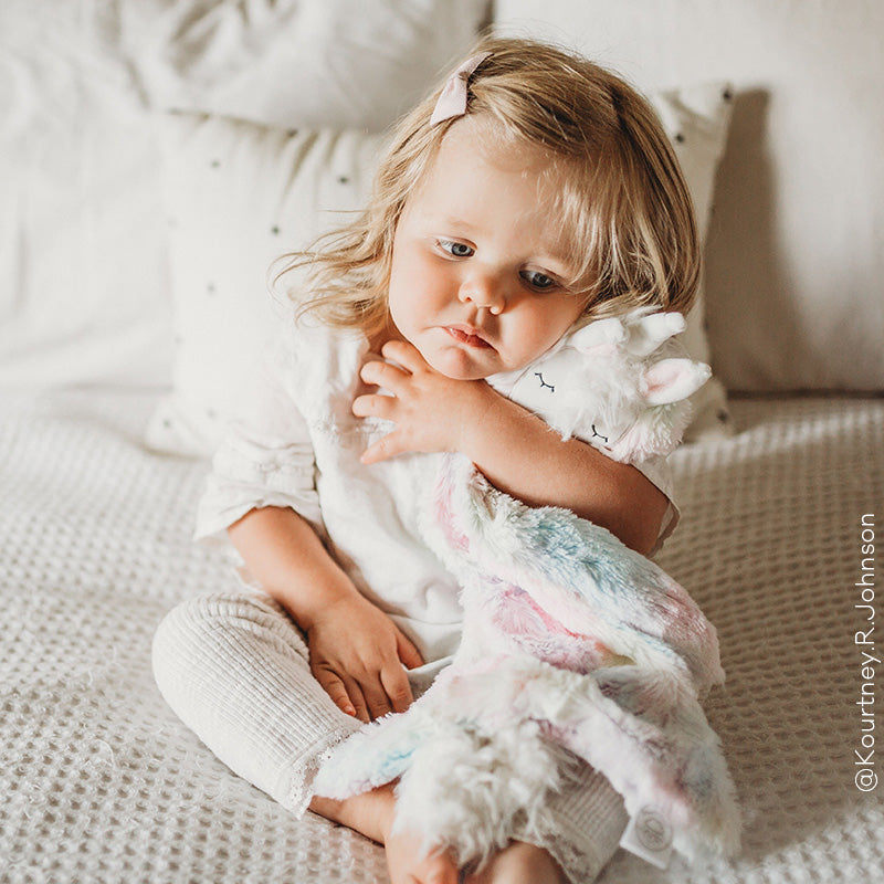 Toddler sitting on a bed hugging a pastel plush lamb, looking thoughtful in a soft neutral bedroom setting
