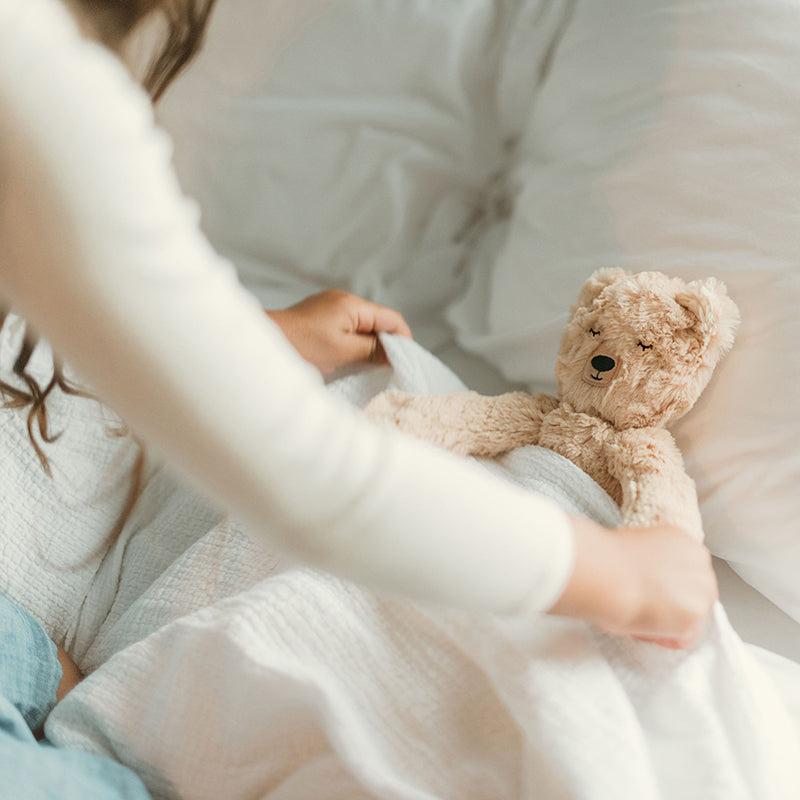 Child tucking a beige plush bear into bed on white quilted bedding