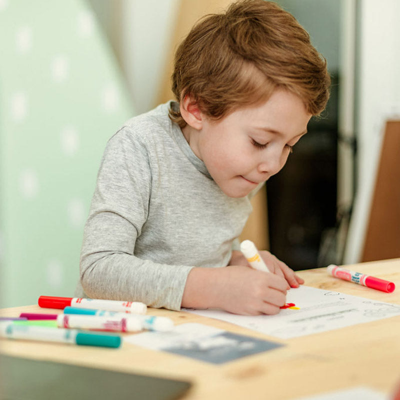 Young child coloring at a table, focused on a worksheet with scattered markers