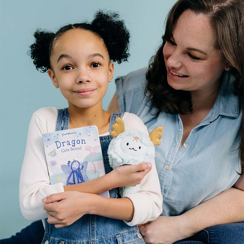 Child holding a Dragon Gets Bored book and plush toy beside an adult