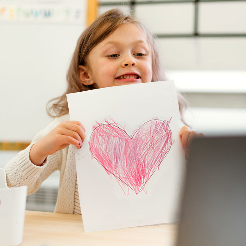 Young girl holding up a paper with a crayon-drawn pink heart in a classroom