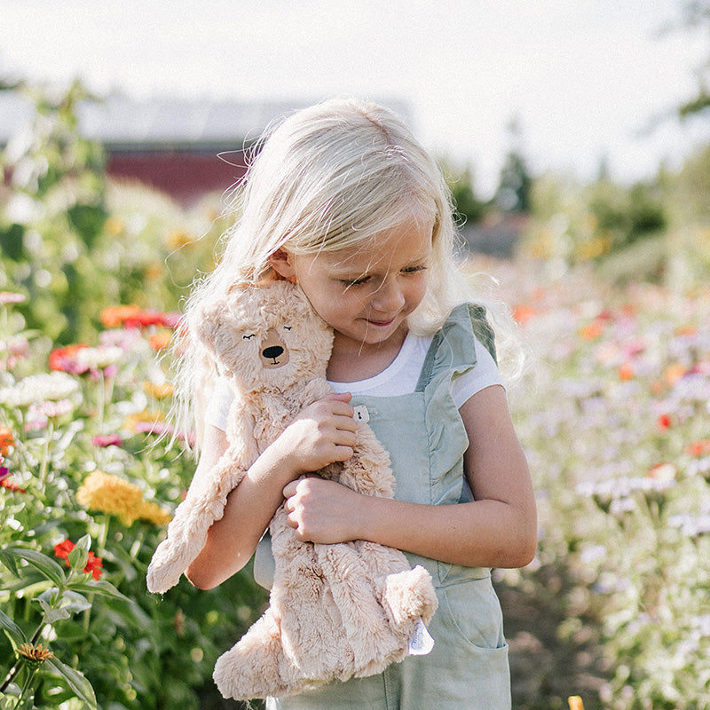 Little girl hugging a beige plush bear while standing in a sunlit wildflower field