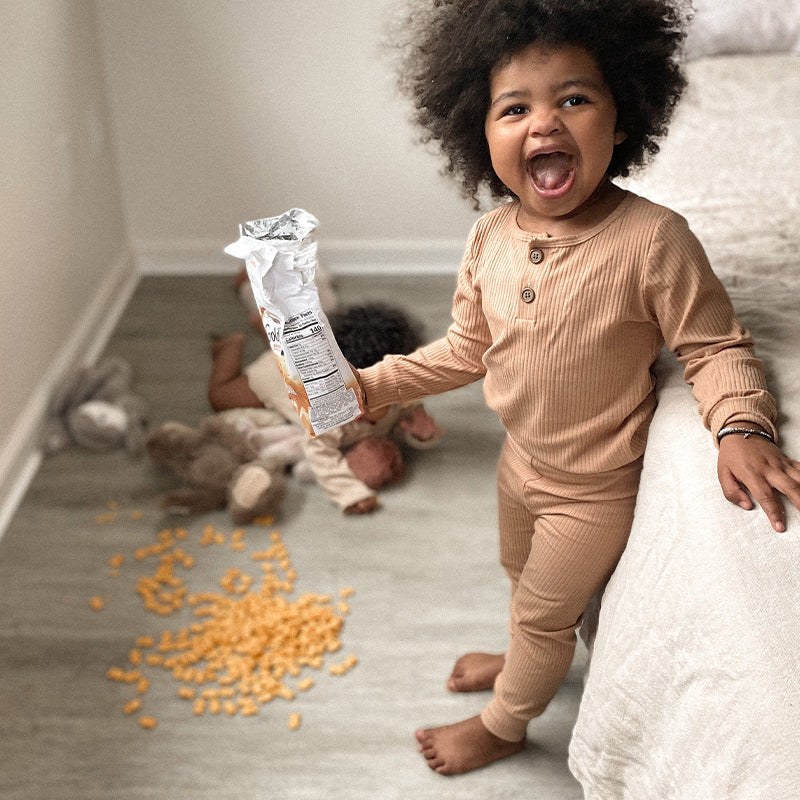 Toddler smiling and holding an open snack bag while cereal is spilled on the floor beside toys in a bedroom