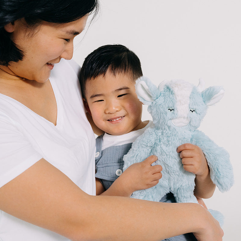 Caregiver holding a smiling child who hugs a blue plush toy with closed eyes against a white background