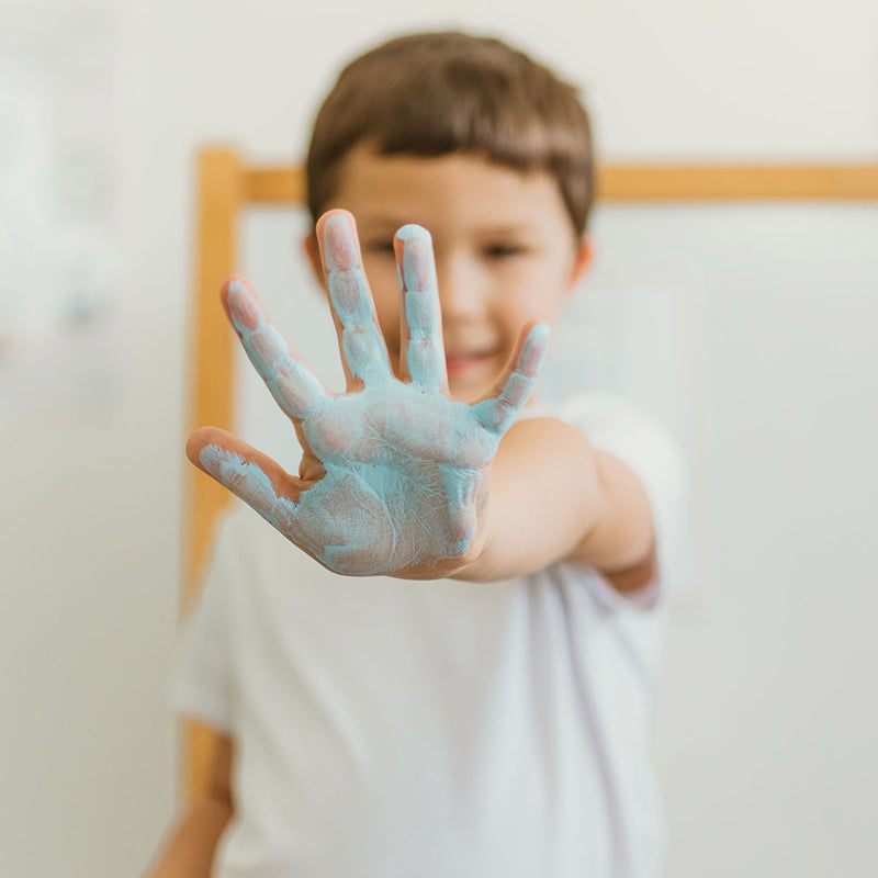 Child holding a blue-painted hand up to the camera in a classroom with a blurred whiteboard behind them