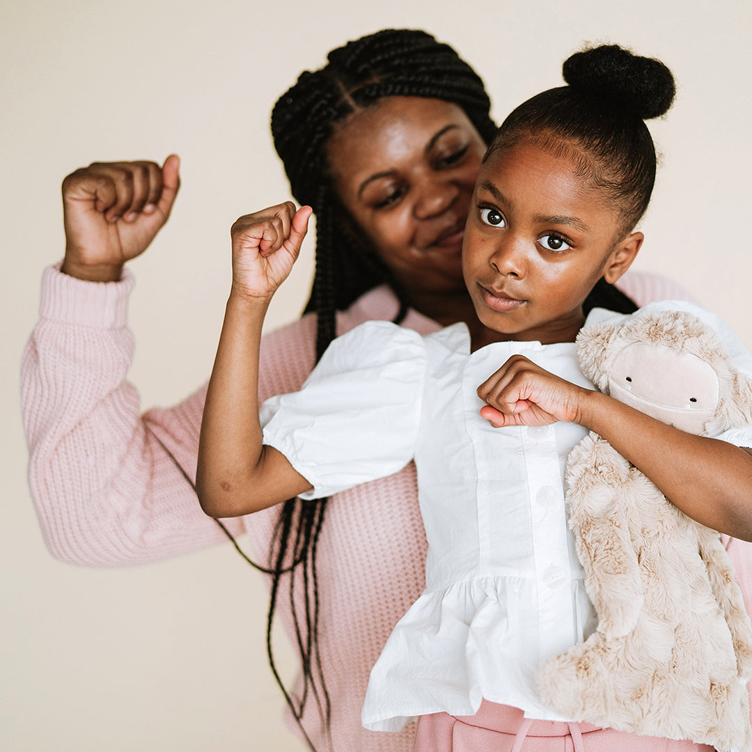 Young girl holding a plush toy and striking a confident pose while a woman holds her from behind against a neutral background