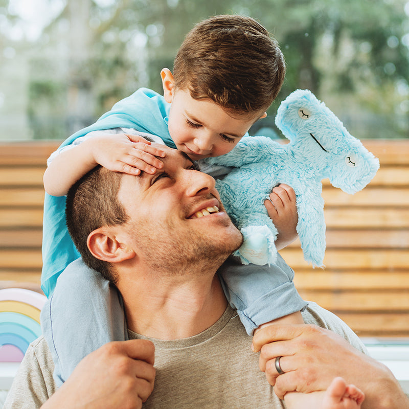 Father smiling with child on his shoulders while the child holds a blue plush toy and gently touches his forehead in a bright room