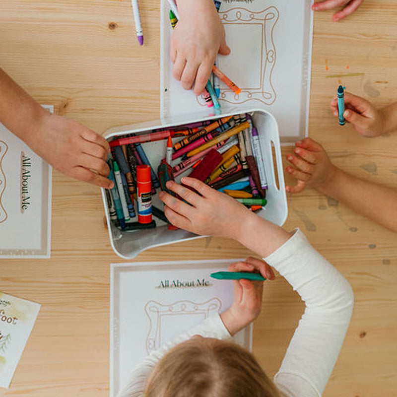 Children sharing crayons and coloring ‘All About Me’ worksheets around a wooden table in a classroom