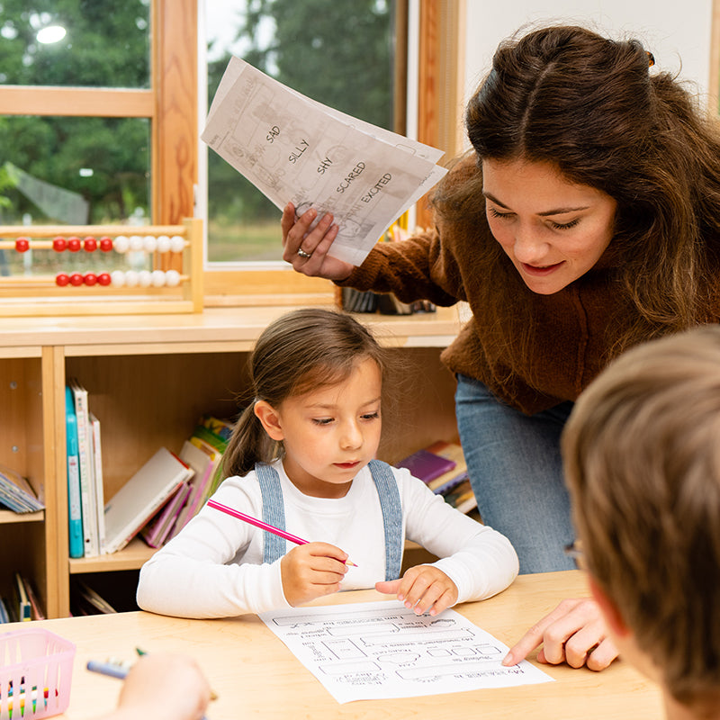 Teacher leaning over to help a young girl complete a worksheet at a classroom table, bookshelves and an abacus visible behind them