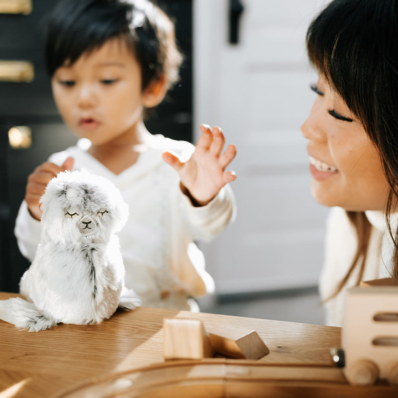 Toddler reaching for a light gray plush toy on a wooden table while a smiling caregiver watches in a sunlit room