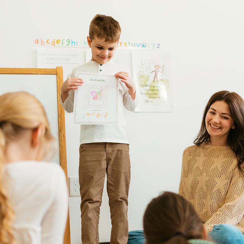 Boy presenting a drawing to classmates while a teacher smiles in a bright classroom